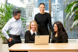 An image of 4 people at a desk smiling looking at a laptop.